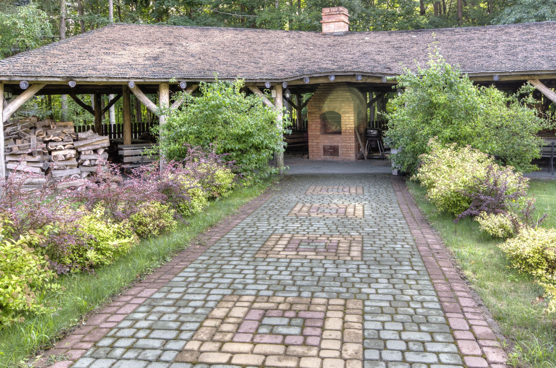 Cobblestone path leading to rustic pavilion with brick oven, framed by shrubs and trees
