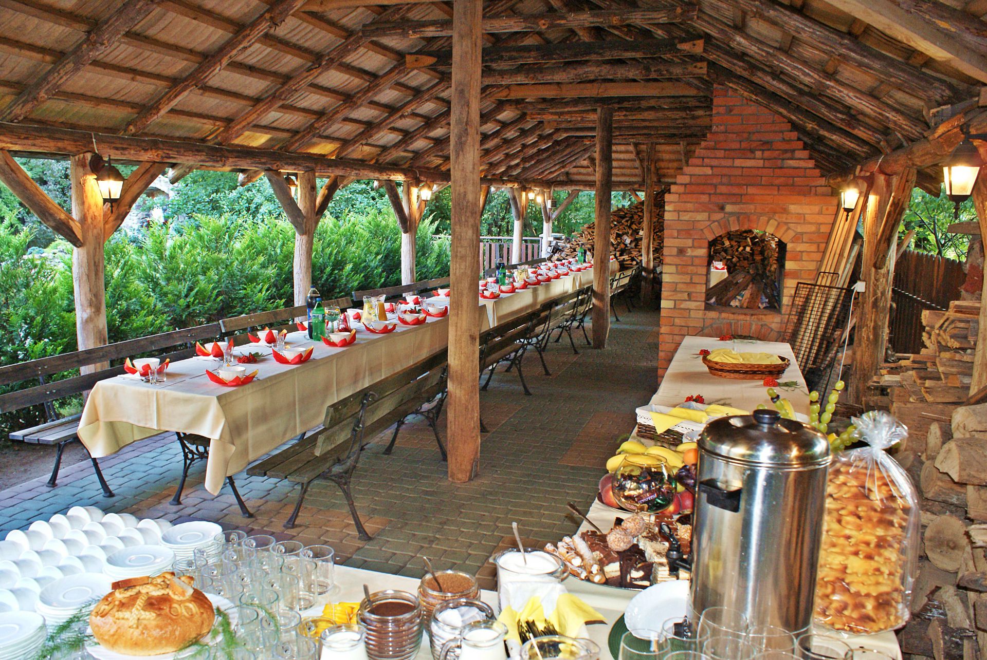 Open wooden dining shelter with long tables set, lanterns, brick fireplace and stacked firewood