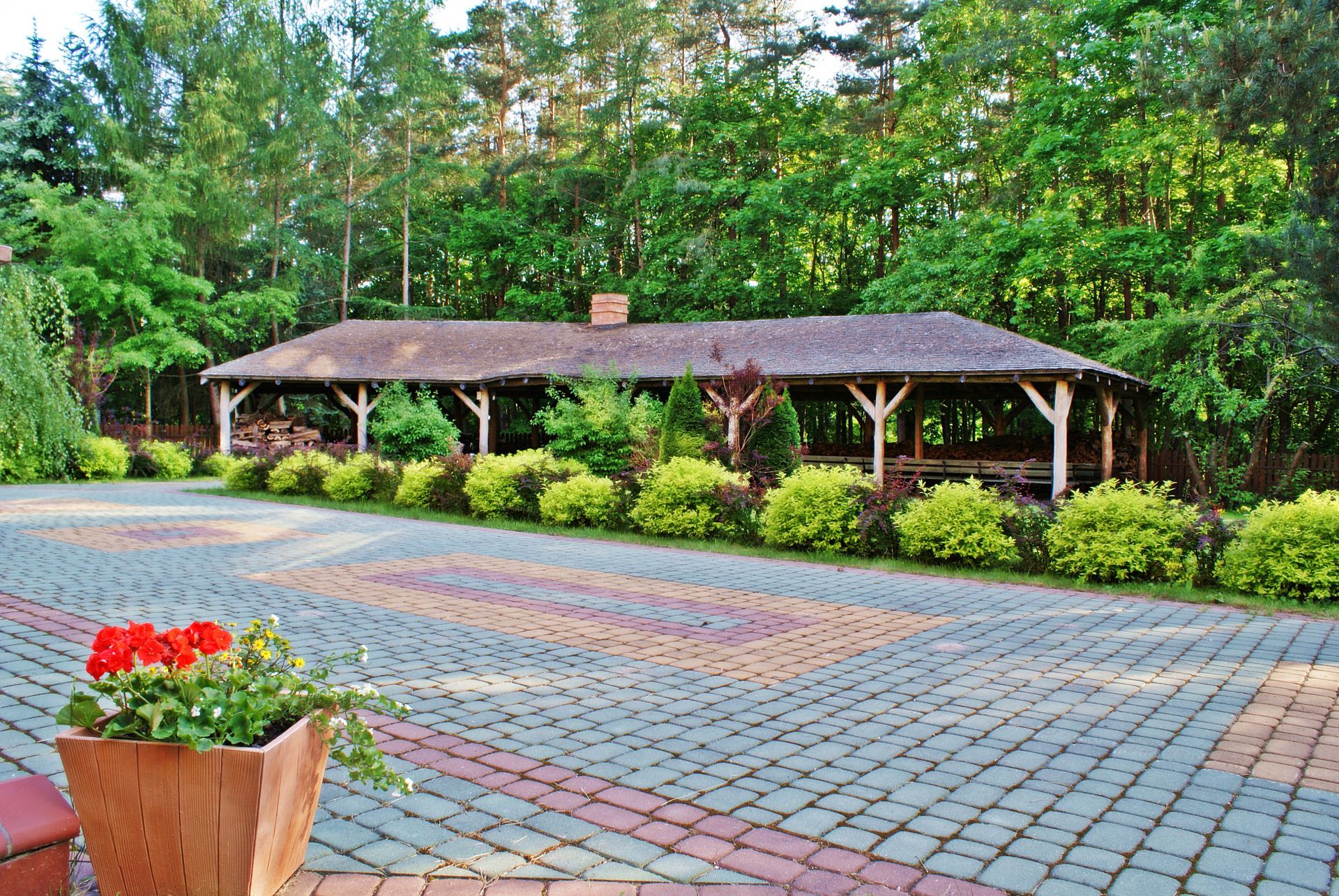 Paved courtyard with planter of red geraniums in foreground and rustic covered pavilion beyond