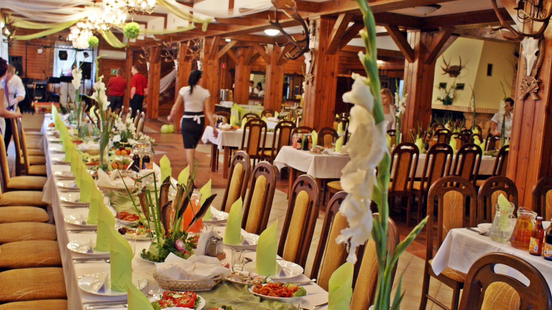 Long banquet table in a wooden hall set with green napkins and floral centerpieces, staff