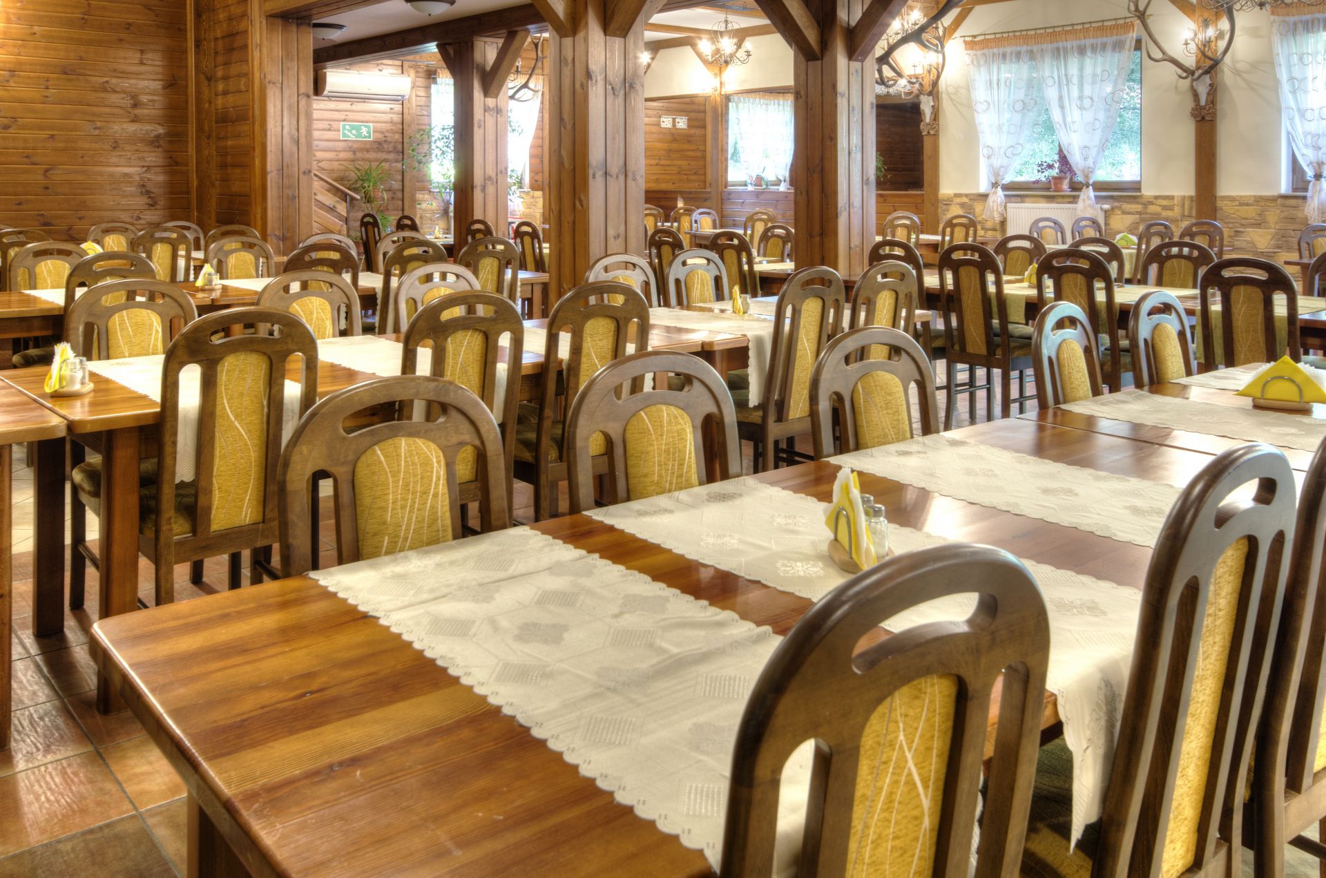 Rustic wooden dining room with many empty chairs and lace runners, warm inviting atmosphere