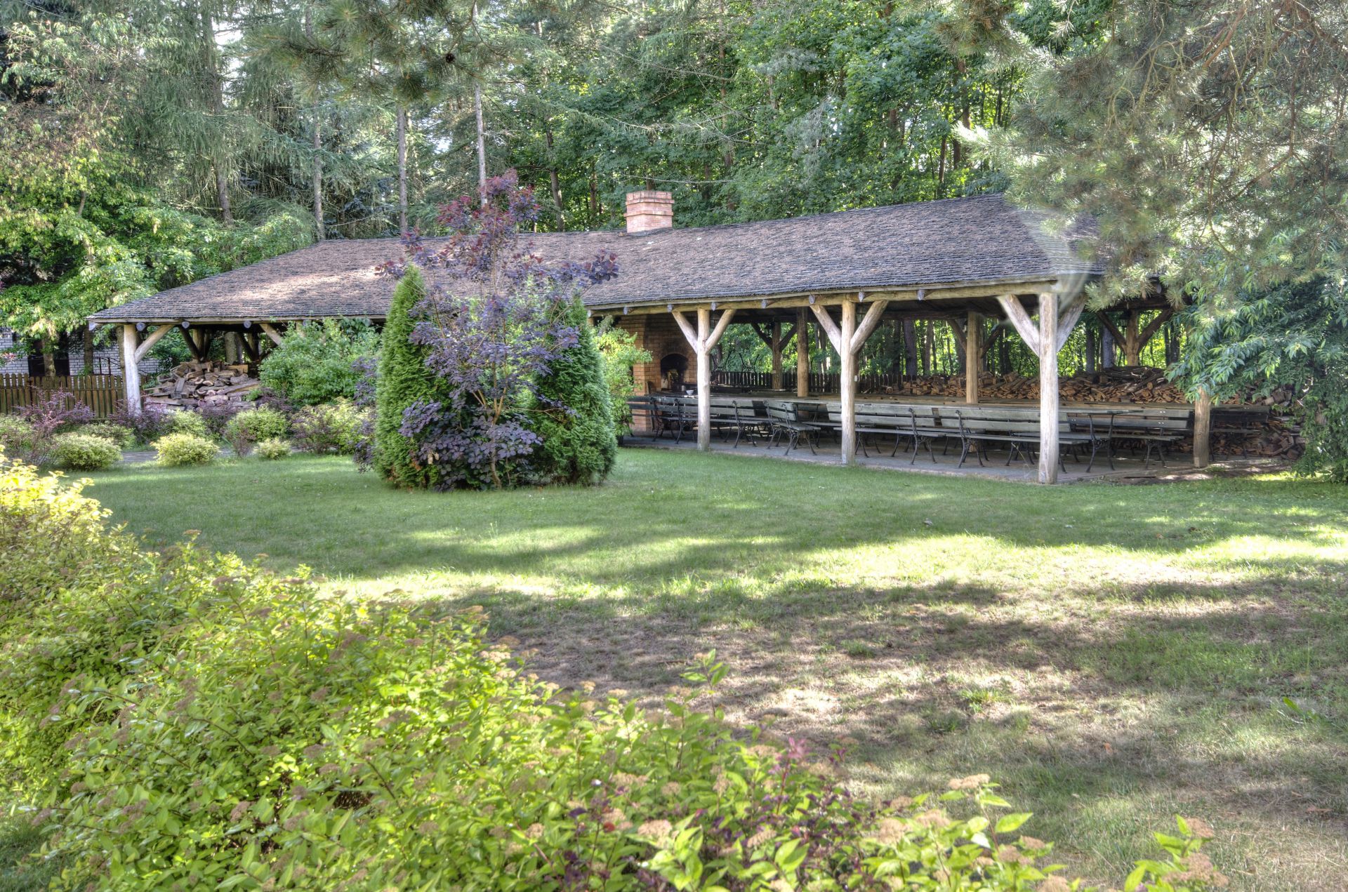 Wooden open pavilion with long benches and stacked firewood in a shady garden.