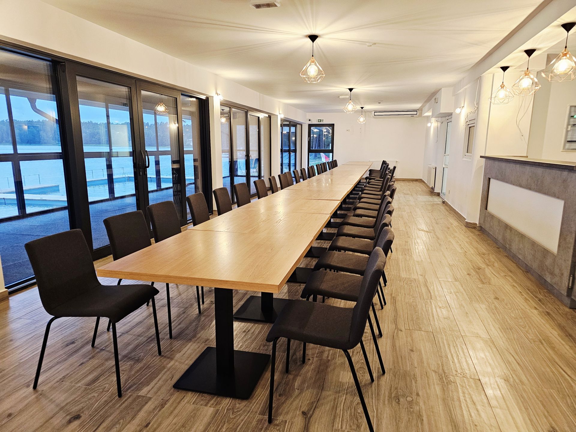 Bright conference space with pendant lights, continuous wooden table and many chairs facing lakeside windows