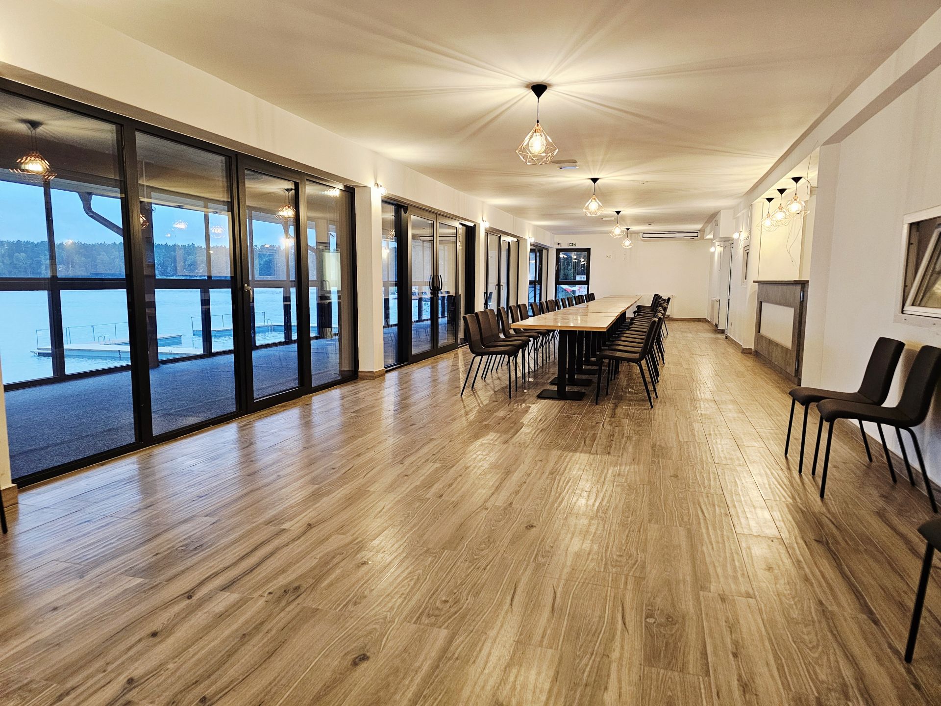 Long meeting room with floor-to-ceiling glass doors, wooden tables and rows of black chairs, lakeside view