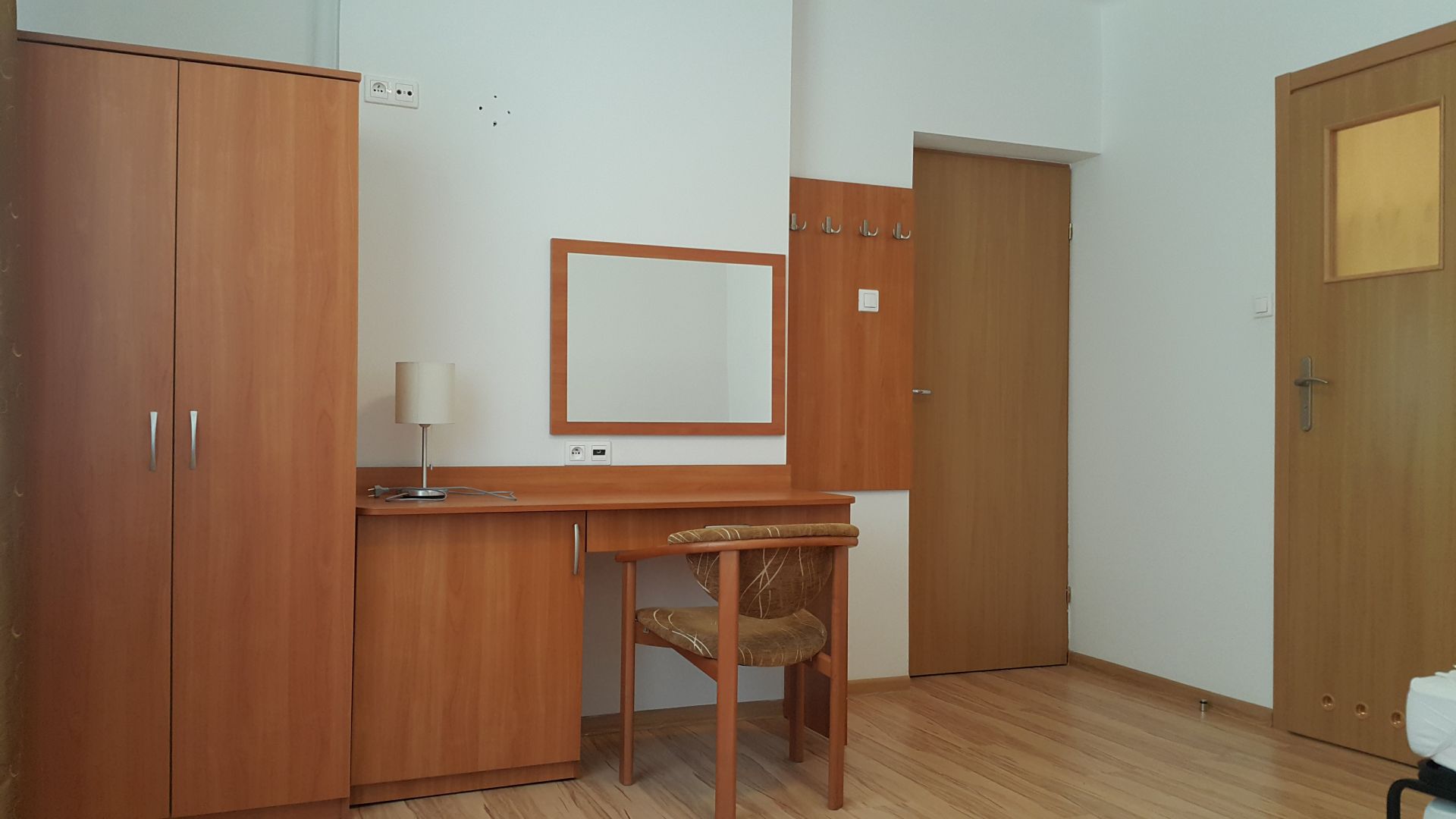 Minimal hotel workspace showing wardrobe, desk with lamp, mirror and wooden chair.