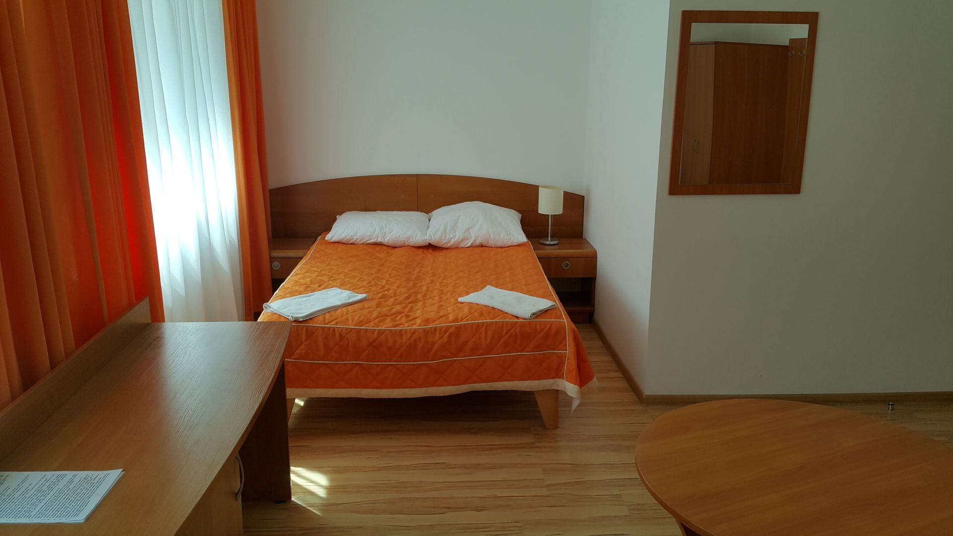 Compact room with double bed, orange quilt, desk and sheer curtained window.