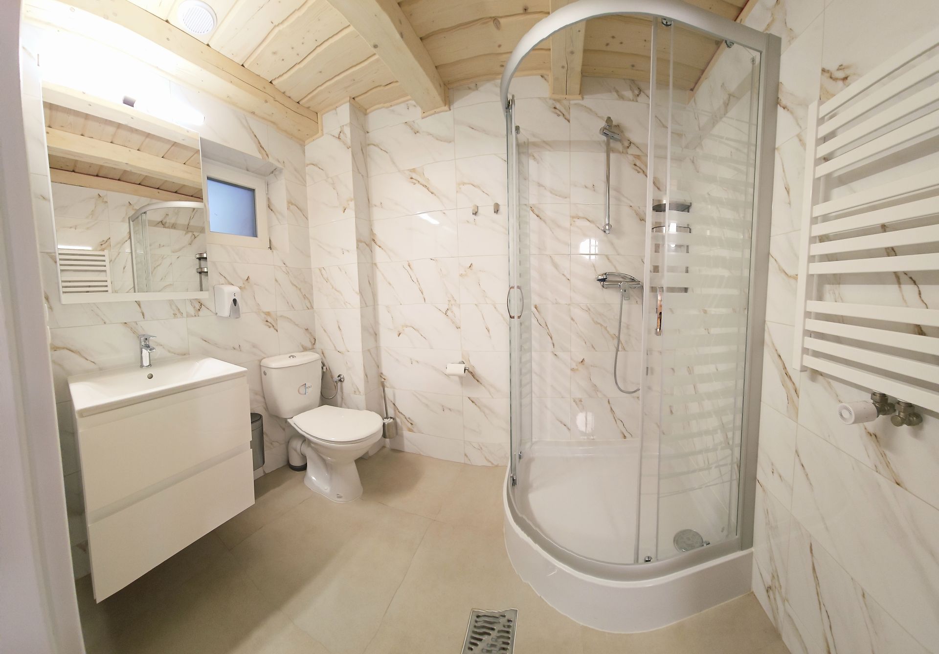 Tiled bathroom with corner glass shower, toilet and vanity under small window.