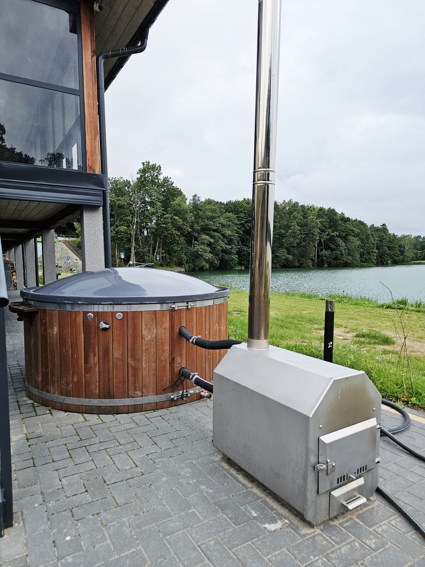 Wooden hot tub with covered lid and stainless heater beside lakeside cabin on overcast day.