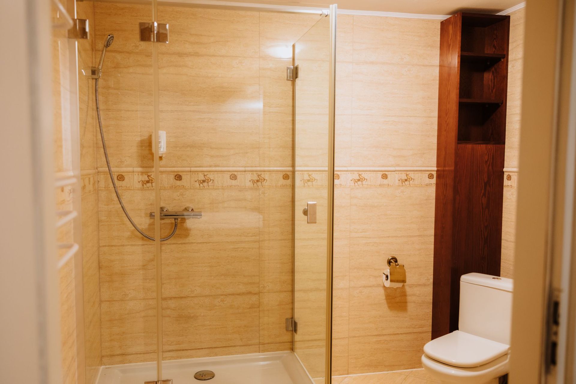 Glass-enclosed shower with handheld showerhead, beige tiles and wooden shelving next to toilet.