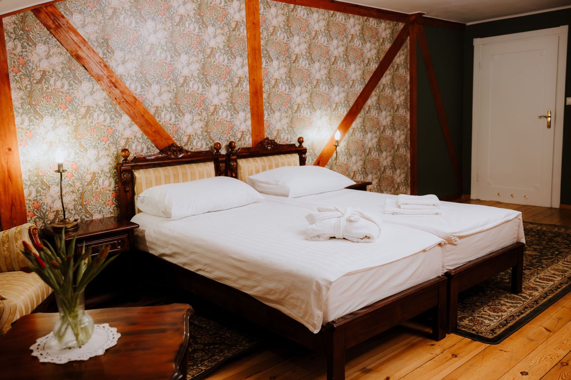 Pair of beds pushed together with ornate headboards, floral wallpaper and patterned floor rugs.