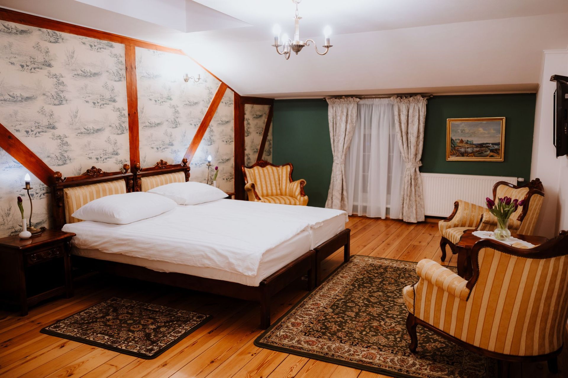 Cozy attic bedroom with carved wooden bed, striped armchairs, green accent wall and lit chandelier.