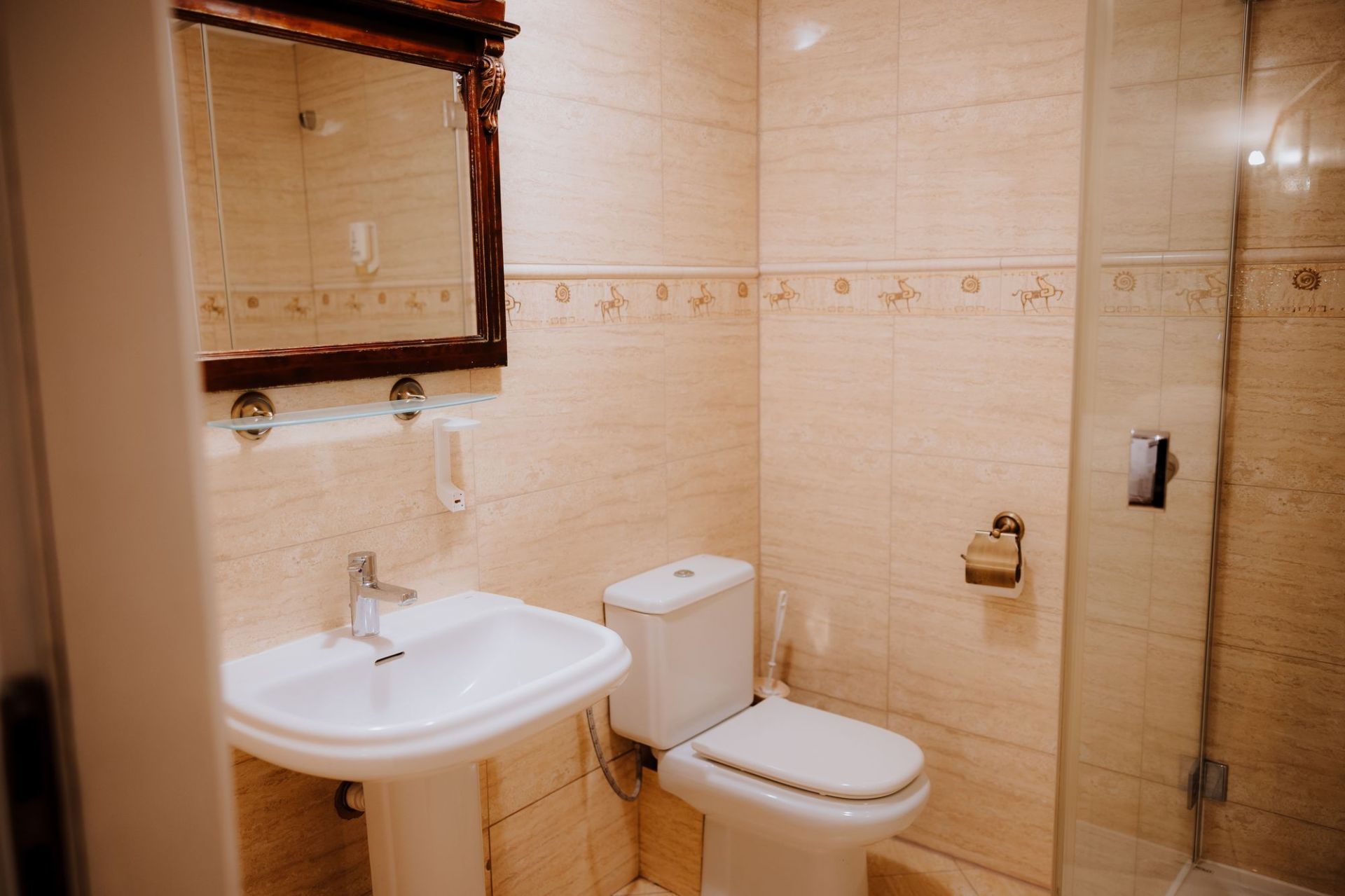 Compact beige-tiled bathroom with white sink, toilet, glass shower door and brass toilet paper holder.